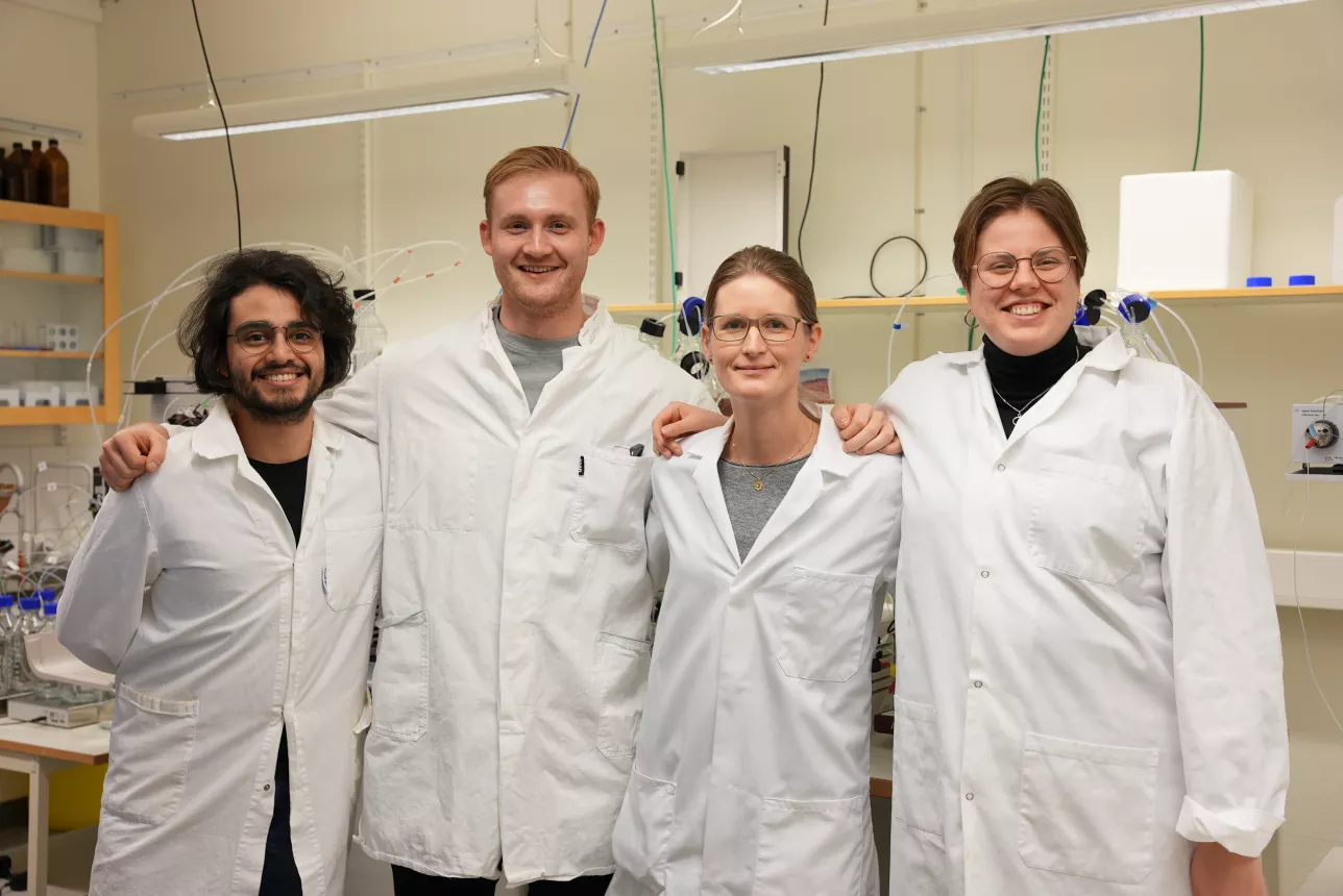 Four researchers in labcoats standing in a lab, smiling to the camera.