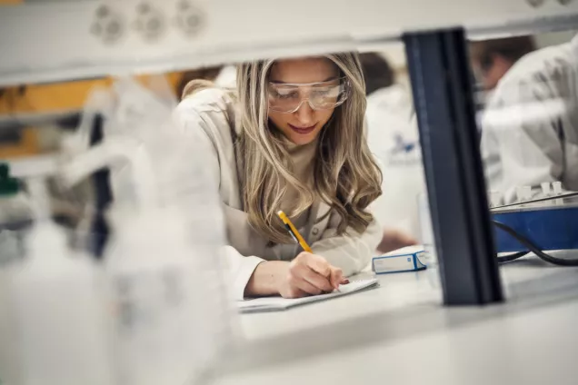 Kvinnlig student sitter i ett labb och skriver i sitt anteckningsblock. Foto.