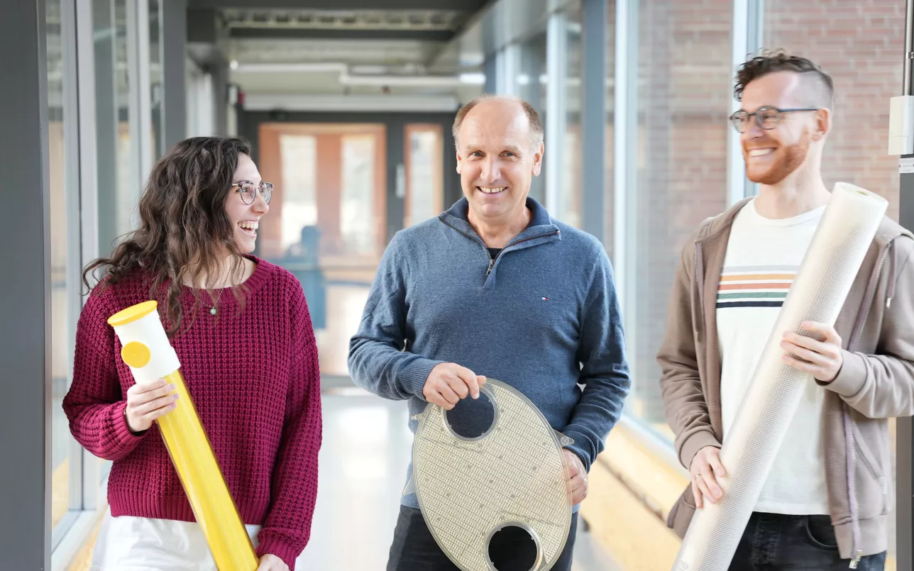 Two men and a woman in a corridor hold up testing equipment. Photo.