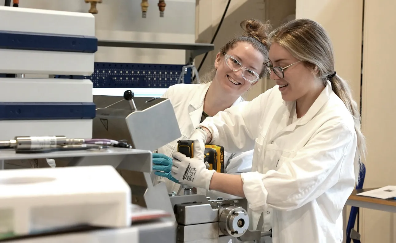 Two happy female employees in lab coats in a big machine hall. Photo.