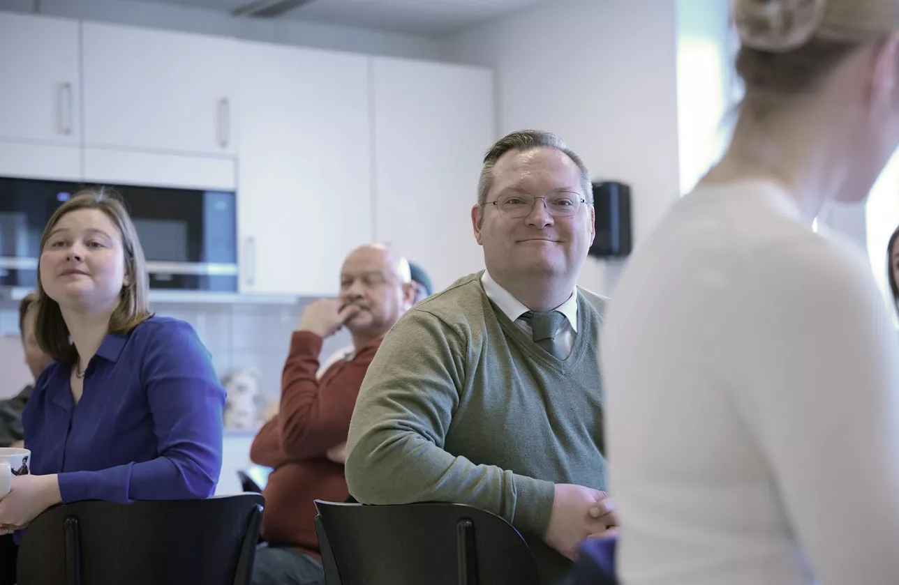A happy male employee looks at the camera in the staff room, surrounded by other employees. Photo.
